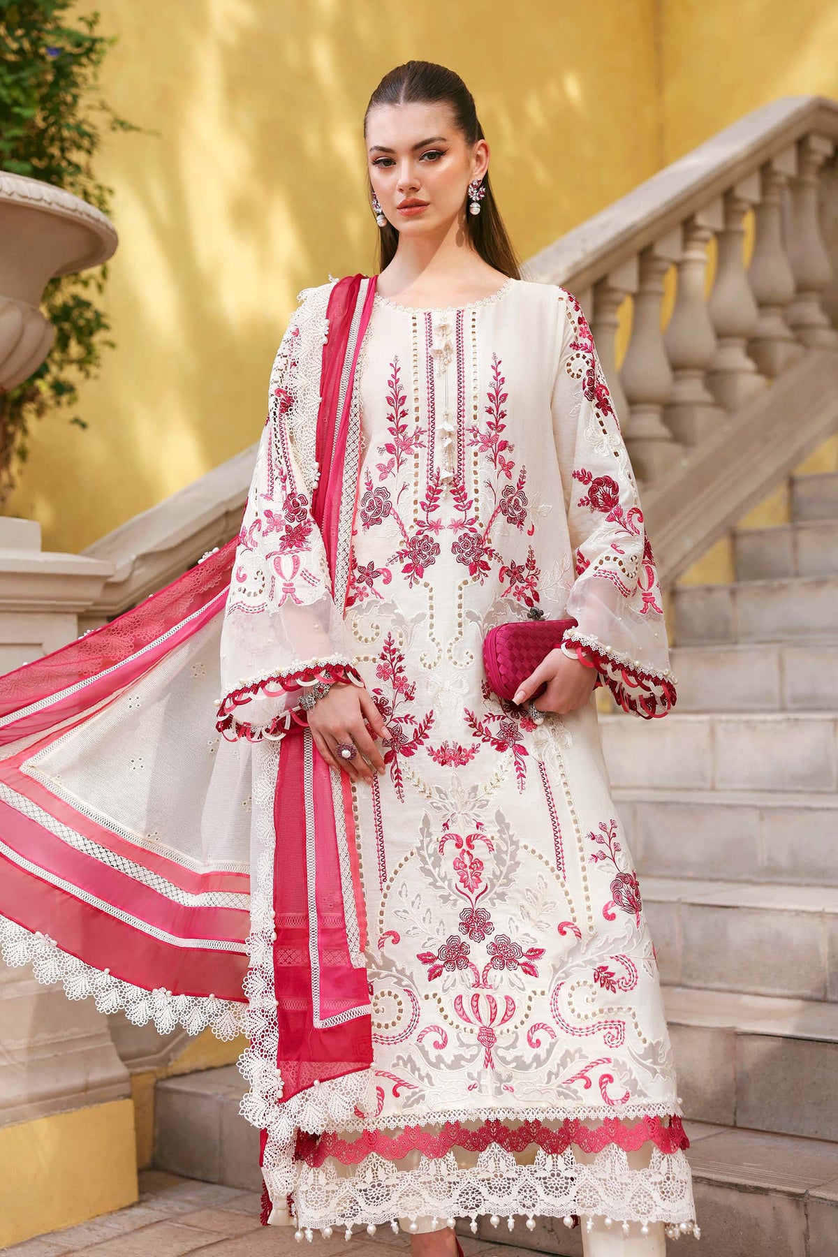 Woman in a white and pink embroidered traditional outfit standing on stone steps with a yellow wall in the background.